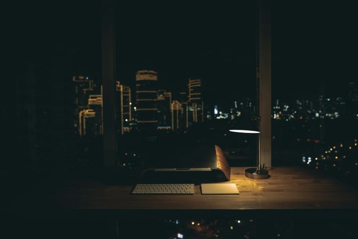 An empty desk lit by a single lamp at night, with city lights through the window